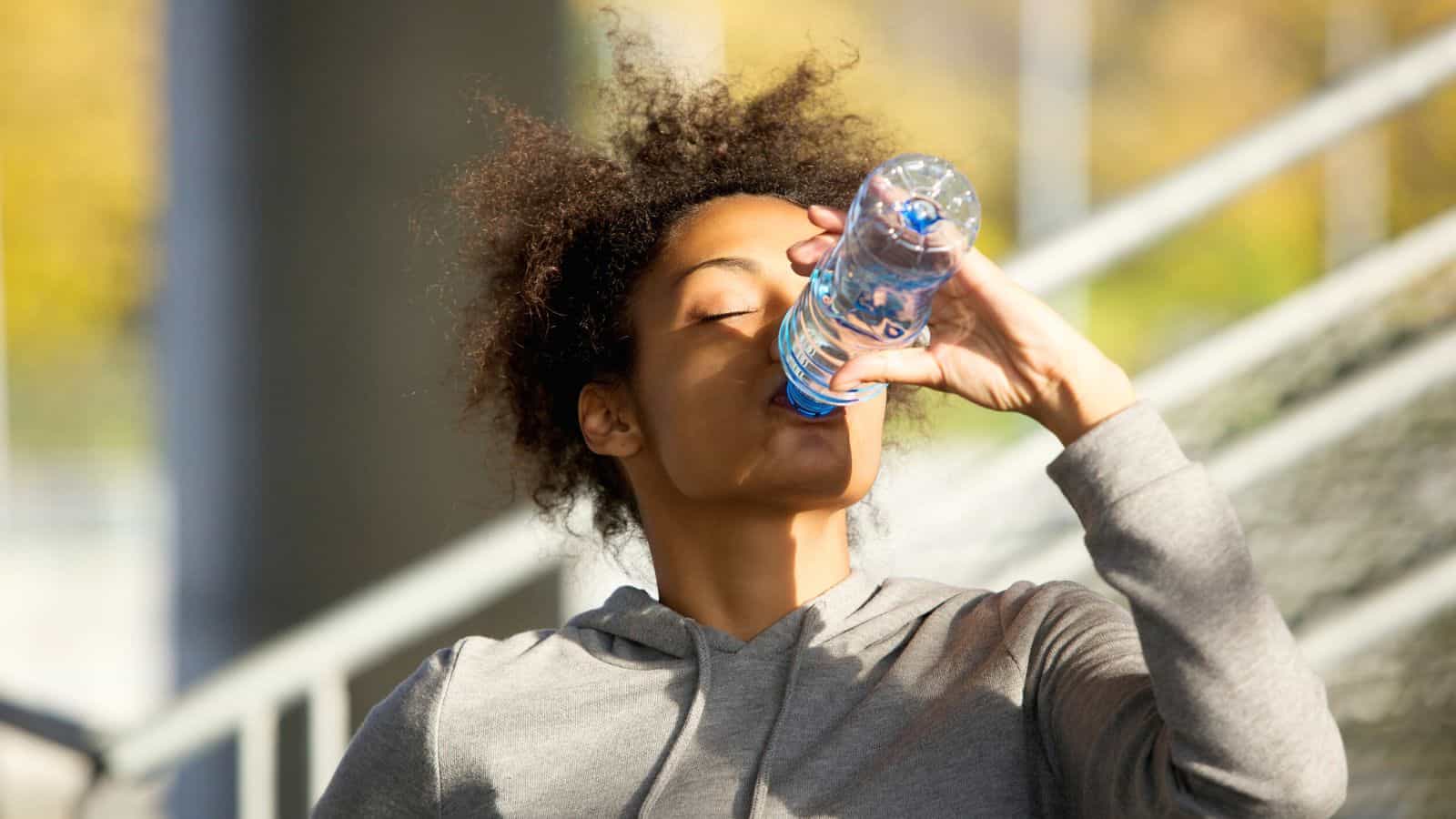 A person in a gray hoodie drinks from a plastic water bottle outdoors. Their eyes are closed, and they are standing in front of a blurred background with a large structure and foliage.