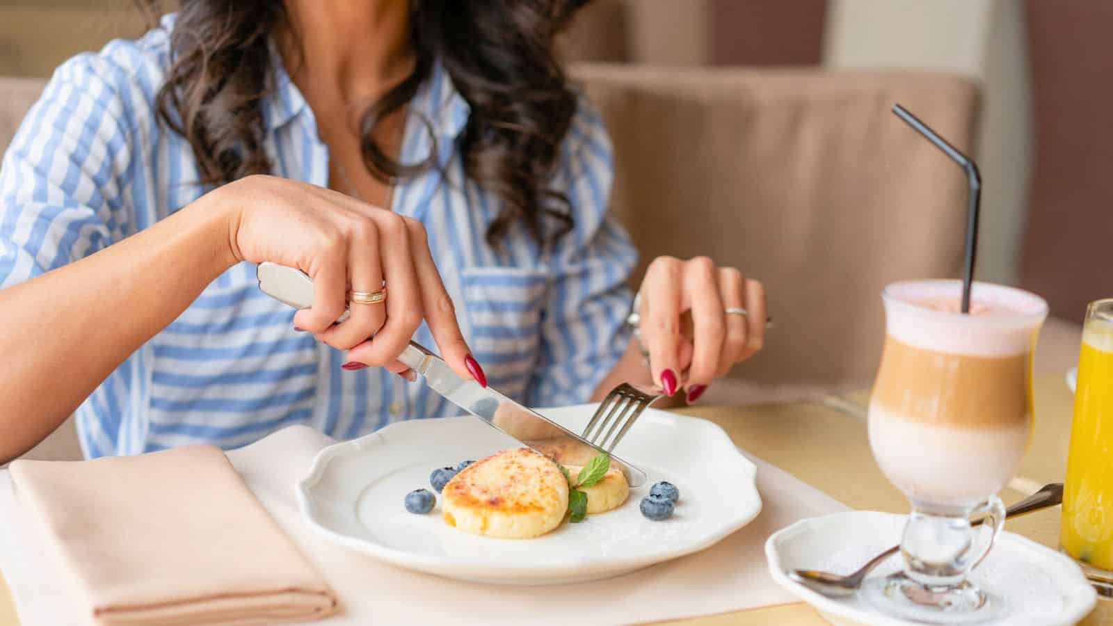 A woman in a blue and white striped shirt is sitting at a table, cutting into a pastry with blueberries on a white plate. There is a latte with a black straw and a glass of orange juice on the table.