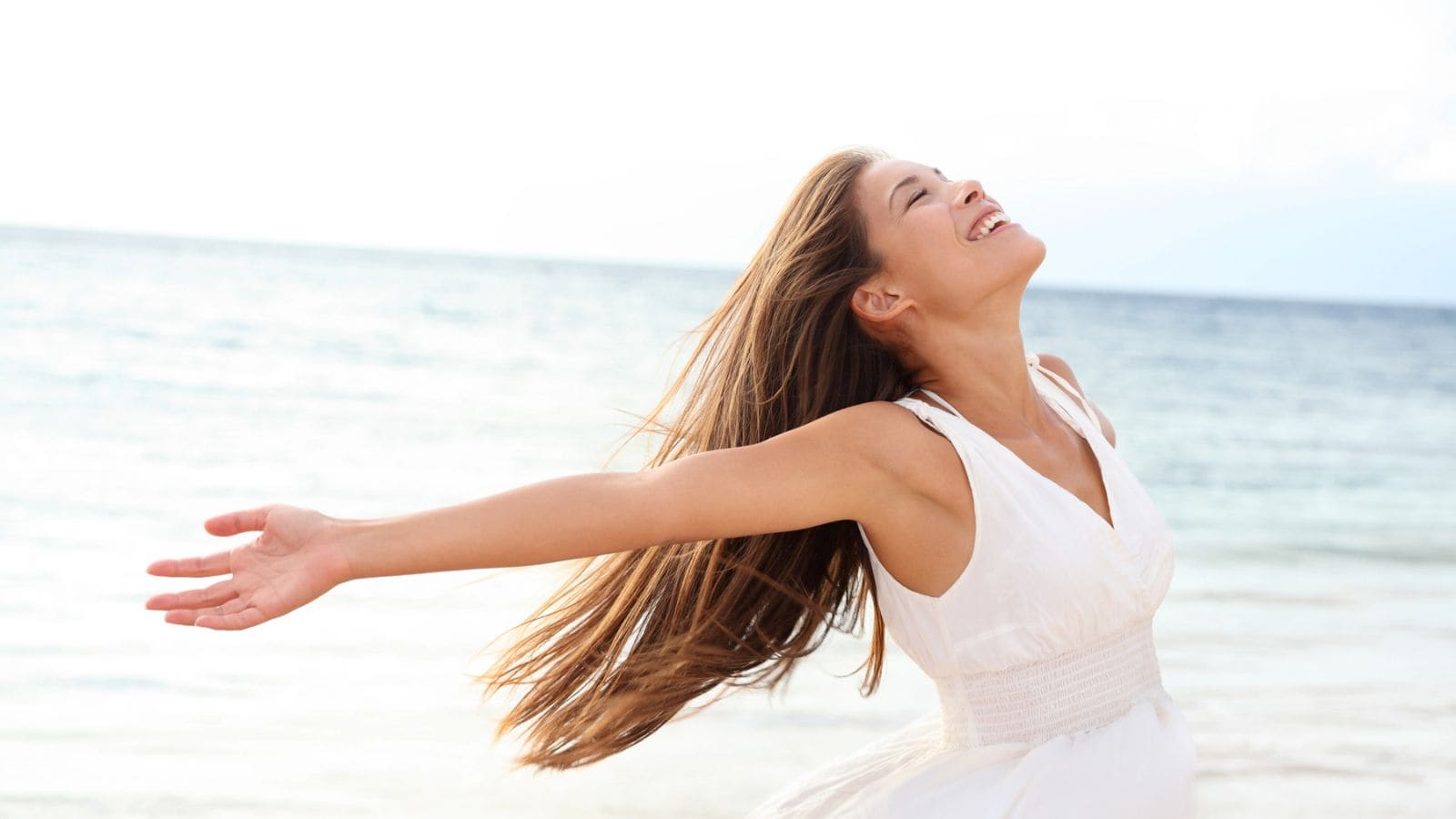 A person with long hair is standing on a beach, wearing a white sleeveless dress. They have their arms outstretched and head tilted back, appearing to enjoy the breeze. The ocean is in the background under a bright sky.