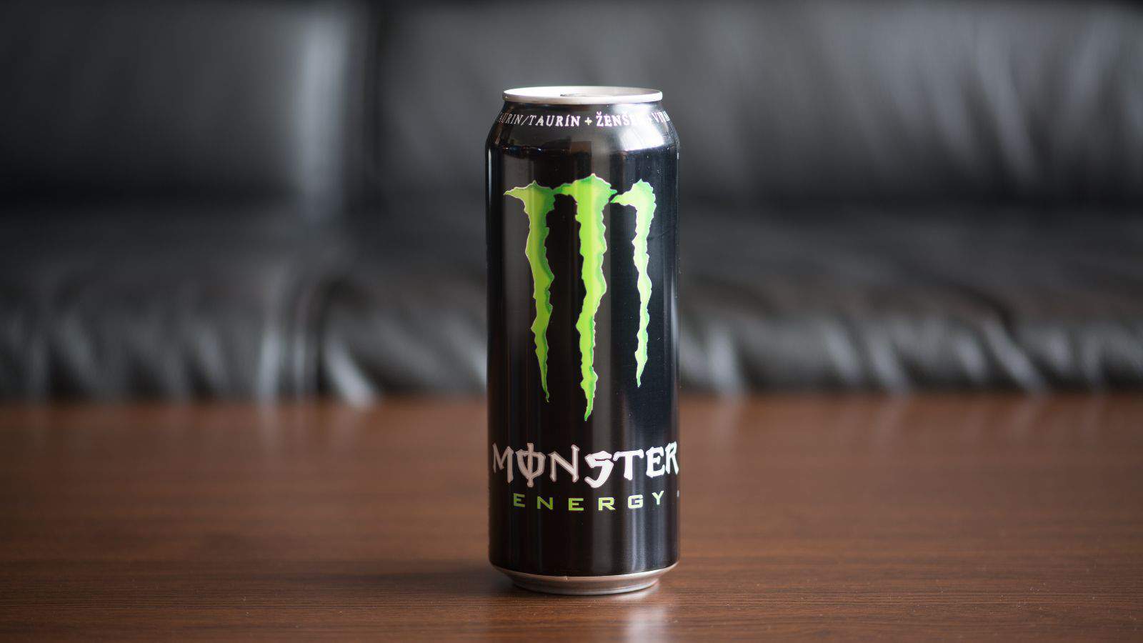 A can of Monster Energy drink sits on a wooden surface, a testament to the allure of processed foods. The black can features the brand's iconic green claw logo and "Monster Energy" text, while in the background, a dark couch remains blurred.