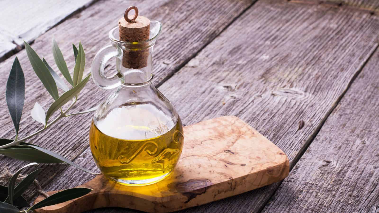 A glass bottle with a cork stopper contains healthy olive oil, sitting on a wooden cutting board. An olive branch with leaves is placed beside it, on a textured wooden surface.