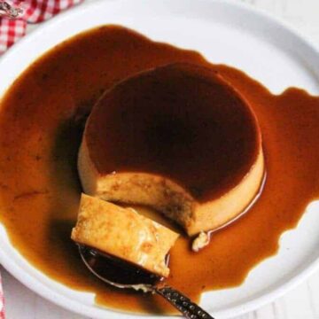 A round serving of flan on a white plate, topped with caramel sauce. A piece of the flan is scooped onto a spoon, and a red and white checkered cloth is partially visible on the side.