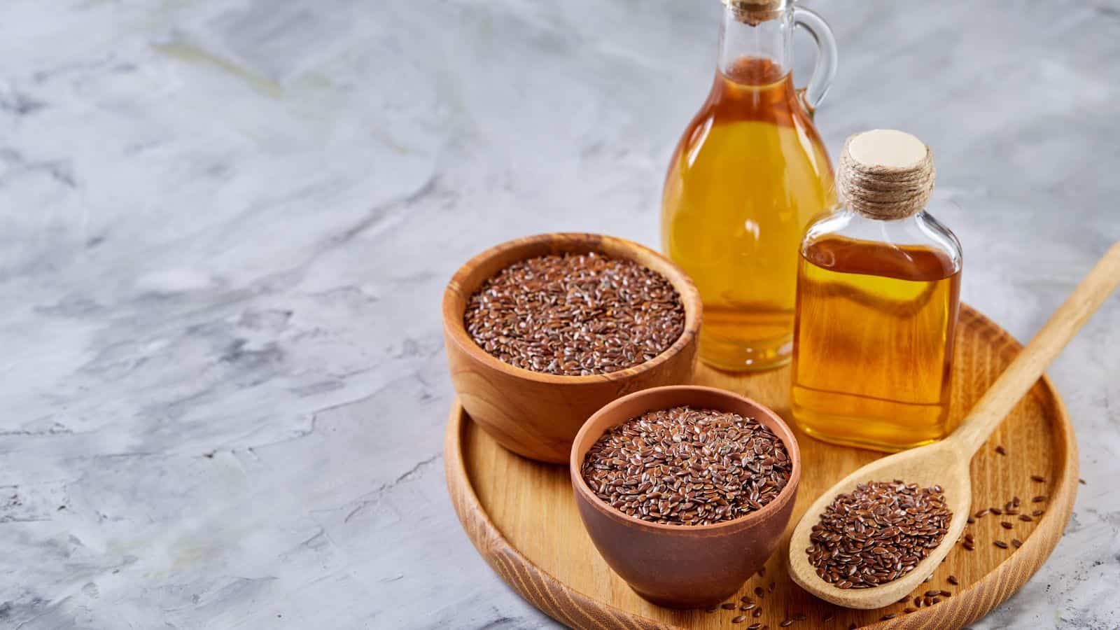 A wooden tray holds two glass bottles of healthy oils and two wooden bowls brimming with flaxseeds. A wooden spoon, dotted with flaxseeds, rests gracefully on the tray. The scene is set against a light, textured background.