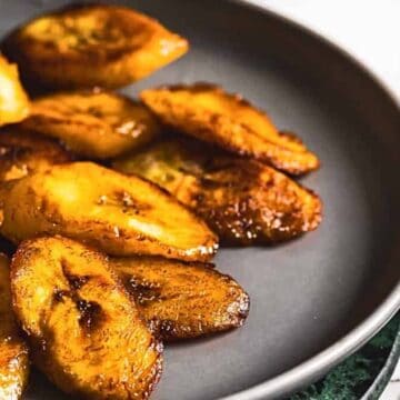 A plate of fried plantains is displayed, featuring golden-brown slices arranged neatly. The plate is placed on a marble countertop, and the plantains have a glistening, slightly caramelized appearance.