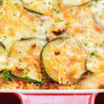 A casserole dish filled with a baked zucchini and cheese dish. Visible ingredients include sliced zucchini, melted cheese, and herbs. The dish is in a red, rectangular baking pan.