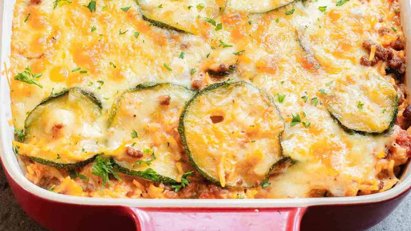 A baked dish in a red casserole dish featuring layers of melted cheese, zucchini slices, and a seasoned meat mixture. The surface is lightly browned, garnished with chopped herbs.