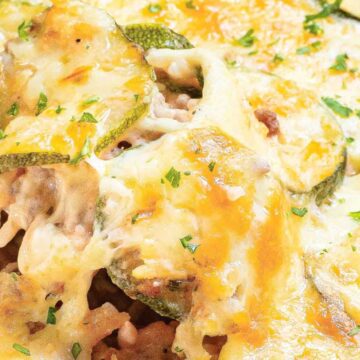 A close-up of a cheesy casserole with a spoon lifting a portion. The dish contains visible layers of melted cheese, sliced zucchini, and a savory filling, garnished with chopped herbs. The meal is baked to a golden brown color.
