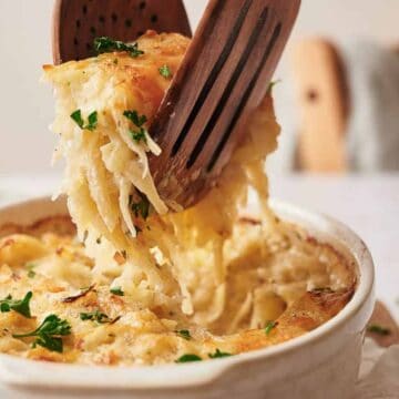 A close-up of a casserole dish with a creamy, cheesy pasta bake. A serving is being lifted with wooden utensils, showcasing the melted cheese and garnished with parsley. A blurred chair and napkin are in the background.