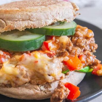 A sandwich on a dark plate featuring minced meat, melted cheese, and red bell peppers, topped with cucumber slices, inside a whole wheat English muffin. Lettuce leaves are visible in the background.