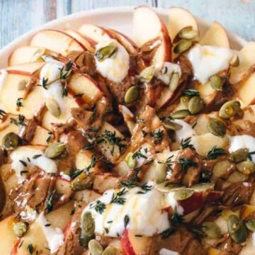 A plate with sliced apples, dollops of yogurt, almond butter drizzle, pumpkin seeds, and fresh thyme on a rustic wooden table.