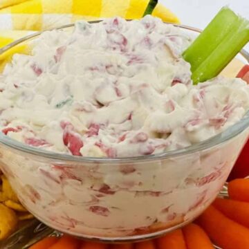 A glass bowl filled with creamy dip containing red bits is placed on a plate. Surrounding the bowl are corn chips, baby carrots, a red pepper, and a celery stick in the dip. A yellow and white checkered cloth is in the background.