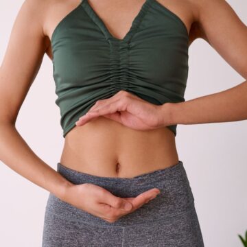 A person standing against a light background is wearing a dark green sleeveless top and gray pants, with arms positioned around the midsection. There are green plants visible in the corner.