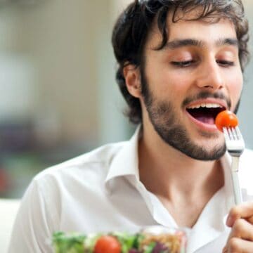 A man with a beard is sitting and holding a fork with a cherry tomato. He is about to eat the tomato. He is wearing a white shirt and there is a bowl of salad in front of him. The background is softly blurred.
