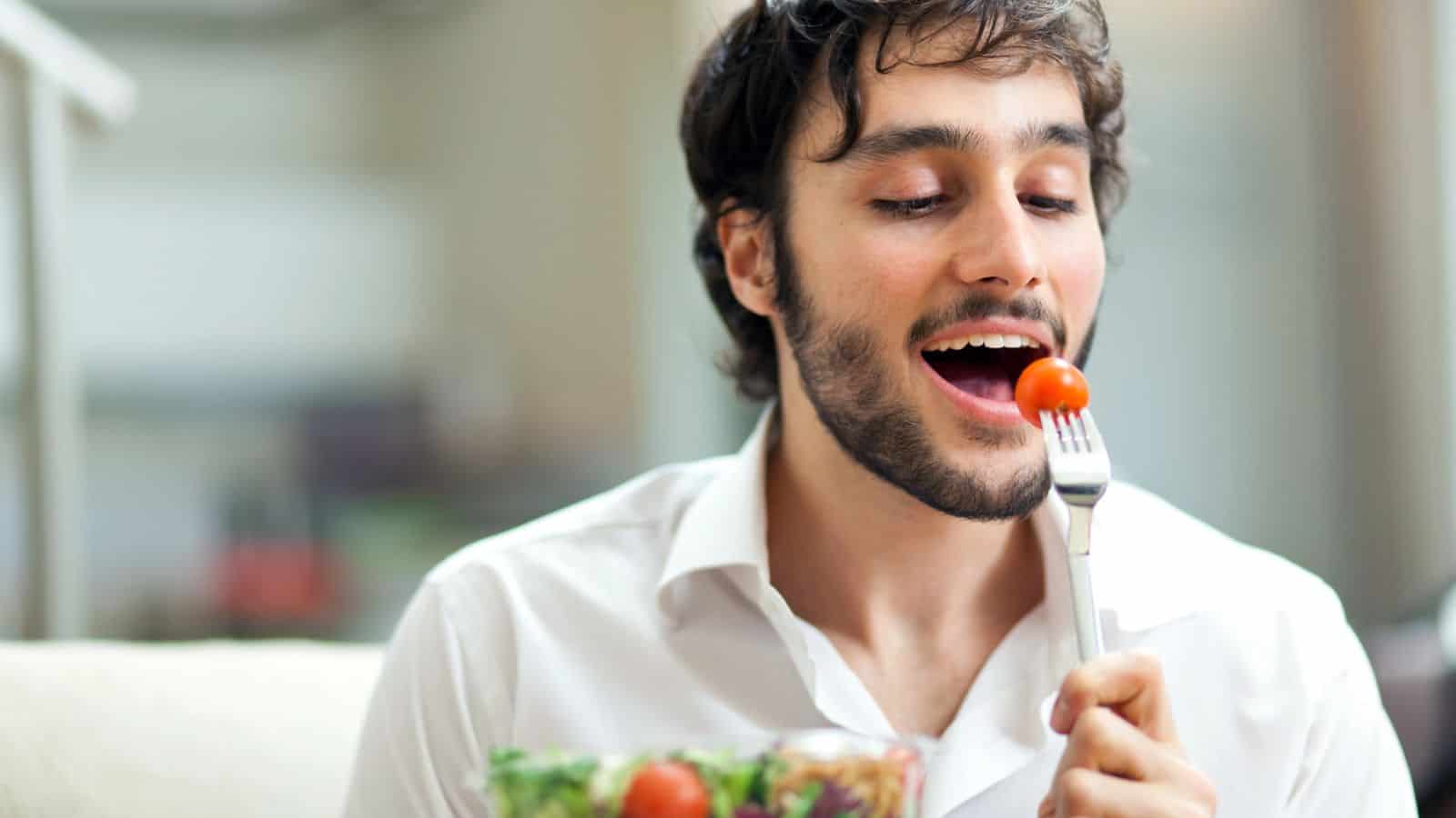 A man with a beard is sitting and holding a fork with a cherry tomato. He is about to eat the tomato. He is wearing a white shirt and there is a bowl of salad in front of him. The background is softly blurred.