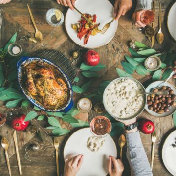A top-down view of a festive table setting with traditional holiday foods like roast chicken, various side dishes, and fruits. Several people are seated around the table, some reaching for food. The table is decorated with greenery and surrounded by plates, glasses, and cutlery.