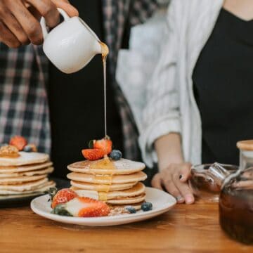 Two people stand by a table with a stack of pancakes topped with strawberries and berries, showcasing surprising Canadian treats. One person pours syrup from a small jug over the pancakes. A glass jar with syrup and a bowl of blueberries are also on the table.