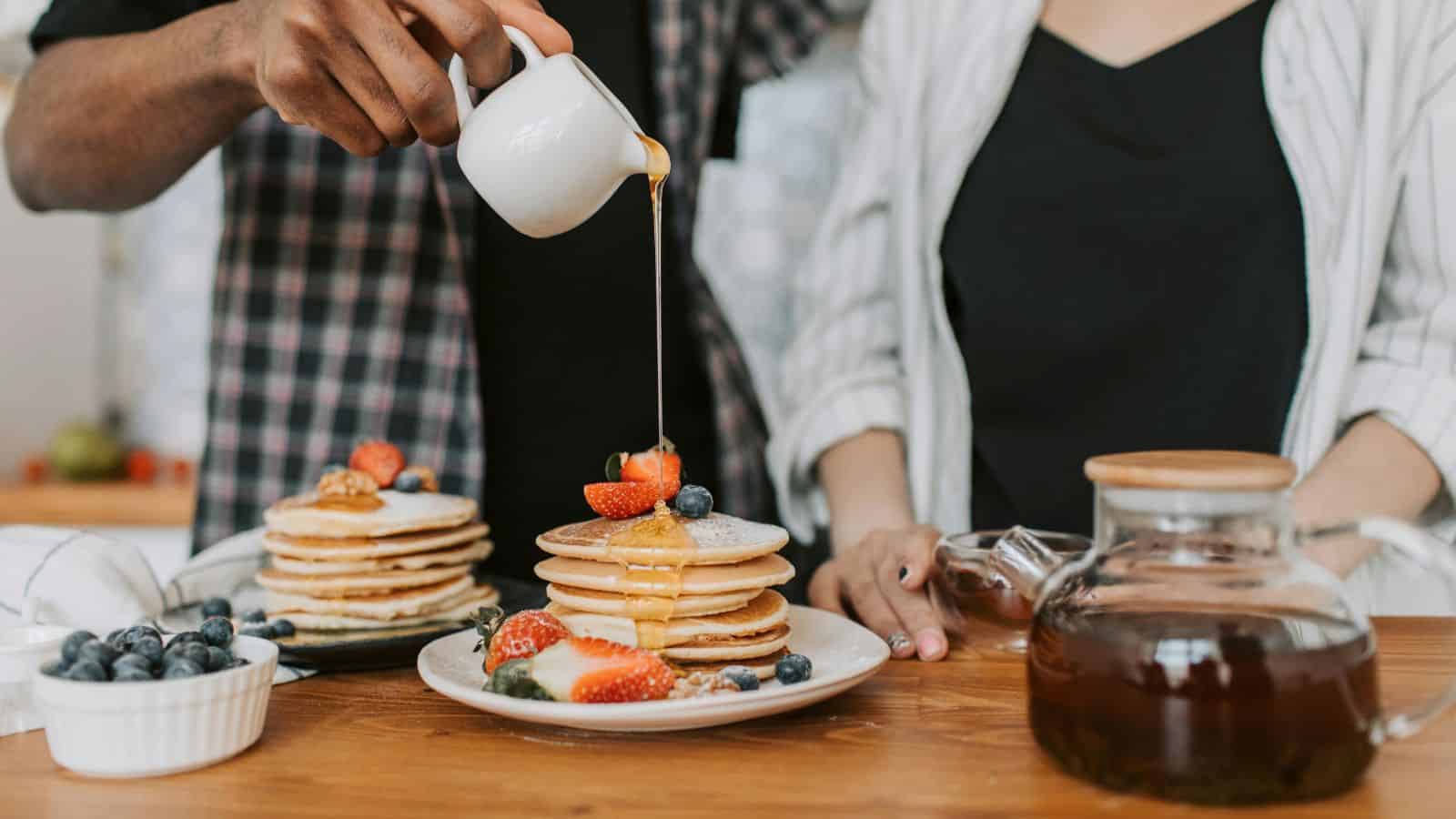 Two people stand by a table with a stack of pancakes topped with strawberries and berries, showcasing surprising Canadian treats. One person pours syrup from a small jug over the pancakes. A glass jar with syrup and a bowl of blueberries are also on the table.