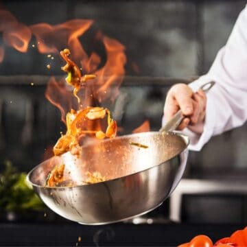 A chef in a white uniform is expertly tossing food in a skillet, showcasing distinctive cooking techniques that cause flames to rise. The background reveals a blurred kitchen filled with various pots and utensils, while red and green vegetables sit prominently on the counter.