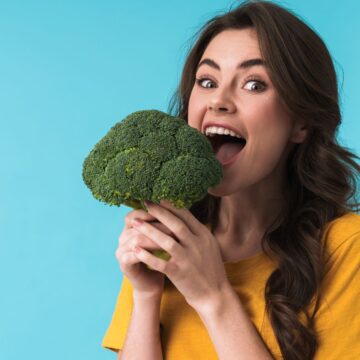 A woman with long brown hair wearing a yellow top holds a large head of broccoli, known for being unexpectedly higher in protein, as if about to eat it. She stands against a blue background.