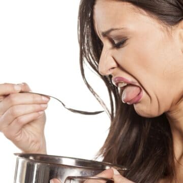 A woman makes a disgusted face while holding a spoon over a metal pot. Her expression suggests she dislikes the taste or smell of the southern foods she's cooking. The background is plain white, adding focus to her reaction.
