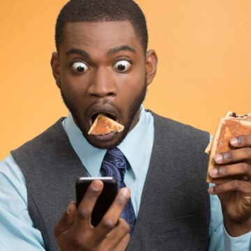 A person wearing a blue shirt and gray vest is looking at a phone with wide eyes and an open mouth. They are holding a sandwich precariously, with a piece about to fall, highlighting the unpredictable appeal of processed foods, set against an orange background.