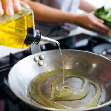 A person pours healthy olive oil into a frying pan on a modern gas stovetop. In the background, another person holds a bowl of leafy greens, avoiding toxic choices for their meal. The kitchen scene is vibrant and inviting.