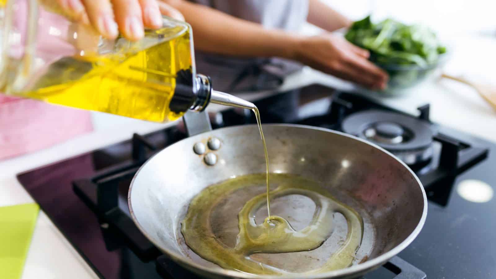 A person pours healthy olive oil into a frying pan on a modern gas stovetop. In the background, another person holds a bowl of leafy greens, avoiding toxic choices for their meal. The kitchen scene is vibrant and inviting.