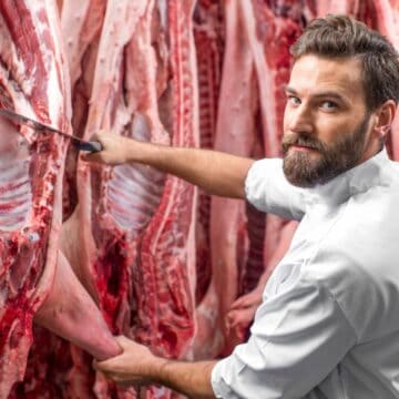 A bearded butcher in a white coat expertly wields a knife to carve different types of meat from a large hanging piece. In the backdrop of this bustling butchery, assorted cuts of meat dangle, showcasing the diverse offerings available in this vibrant shop.