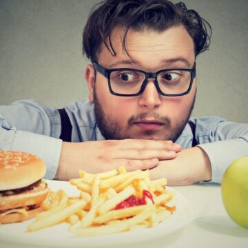 A man with glasses looks intently at a cheeseburger and fries on a plate, with an apple sitting separately. As he contemplates his choice, he wonders if his body's craving for something simple like an apple is how your body asks you to cut back on salt.