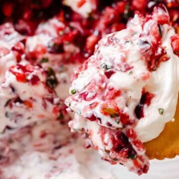 A close-up of a cracker being dipped into a creamy spread mixed with chopped cranberries and herbs. The vibrant red of the cranberries contrasts with the white cream, offering a textured appearance.