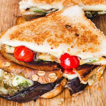 A grilled sandwich, insanely delicious, cut in half on a wooden board, brimming with eggplant, melted cheese, zucchini, and cherry tomatoes. A vintage knife rests beside it while a small bowl of green sauce adds to the allure of this delicious dish.