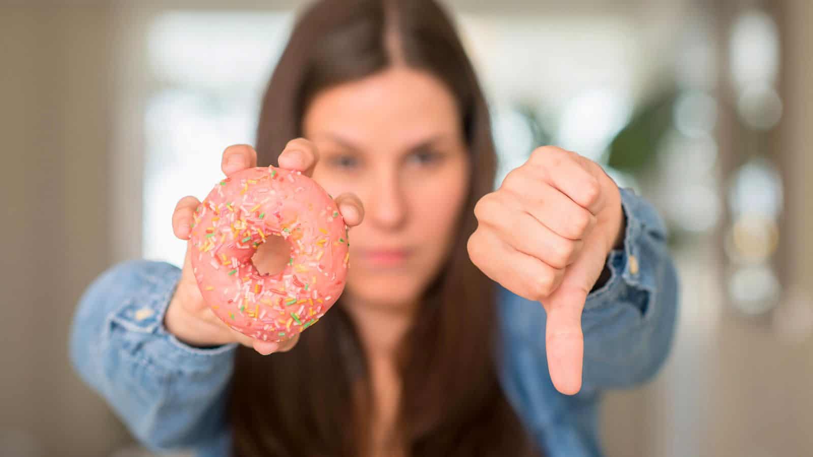 A woman holds a pink frosted donut with sprinkles in one hand while giving a thumbs down with the other hand. She is wearing a denim jacket and has long brown hair. The background is blurred.