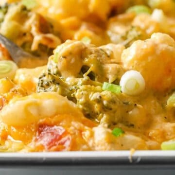 A close-up of a baked casserole dish containing a cheesy vegetable medley. Broccoli, cauliflower, and bell peppers are visible, topped with melted cheese and sliced green onions. A serving spoon rests in the dish.