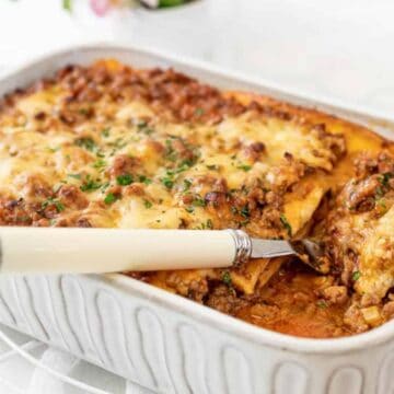 A casserole dish holds freshly baked lasagna topped with melted cheese and garnished with parsley. A serving spatula is lifting a portion from the corner, revealing layers of pasta, meat sauce, and cheese.