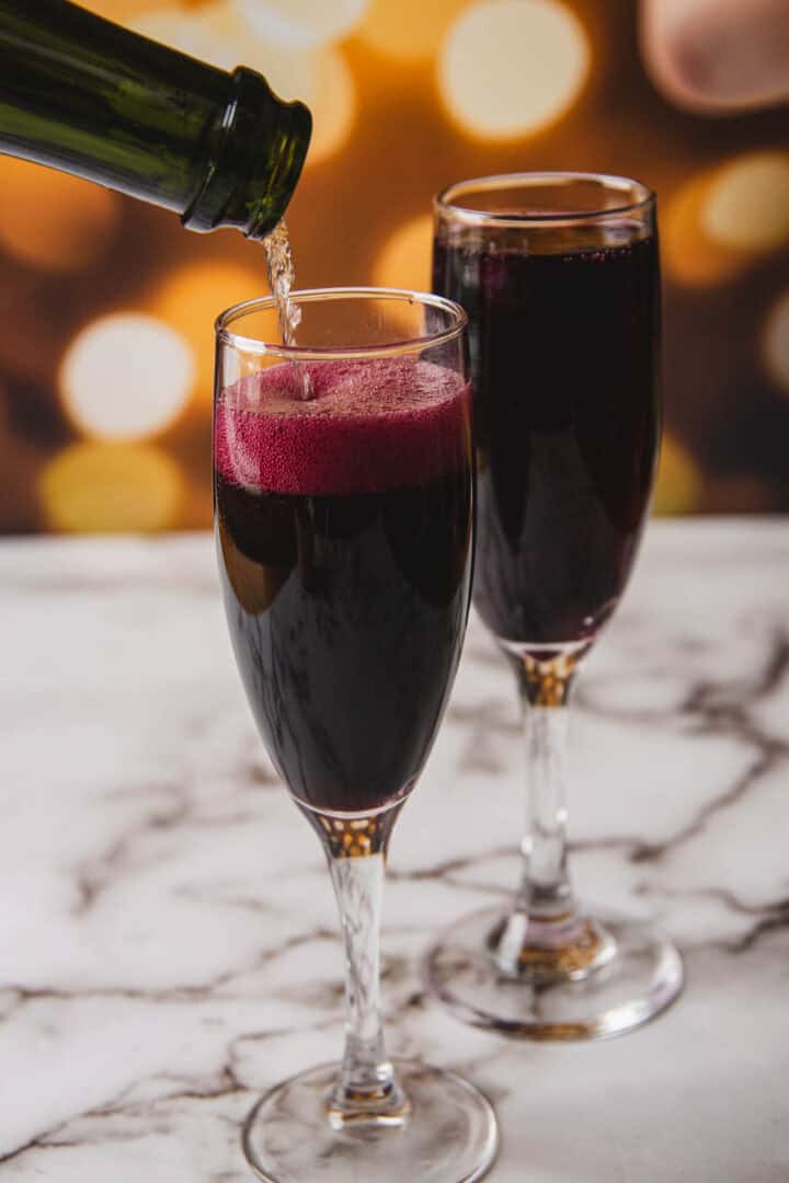 Two champagne flutes filled with a dark red liquid are on a marble surface, reminiscent of a classic Kir Royale. One glass is being topped up from a green bottle, capturing the essence of the cocktail. The background features a bokeh effect with warm tones.