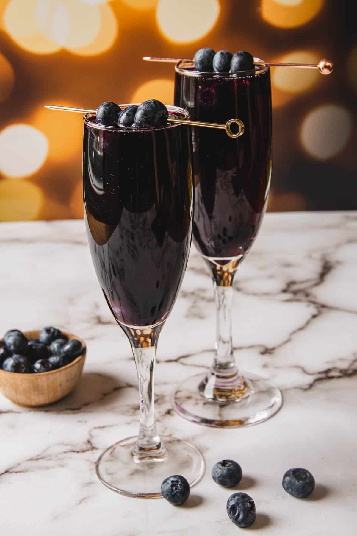 Two tall glasses filled with a dark red drink, resembling a Kir Royale, each garnished with skewered blueberries, sit on a marble surface. A small wooden bowl and scattered blueberries are in the foreground, set against a blurred, warm-toned background.