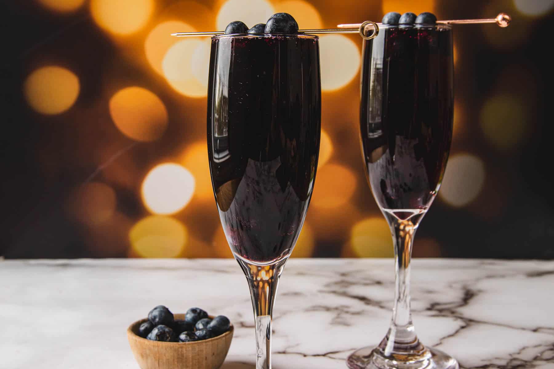 Two champagne flutes filled with a dark red beverage, resembling the classic Kir Royale, are garnished with blueberries on skewers. A small wooden bowl containing blueberries rests on a marble surface, while soft, warm bokeh lights illuminate the background.