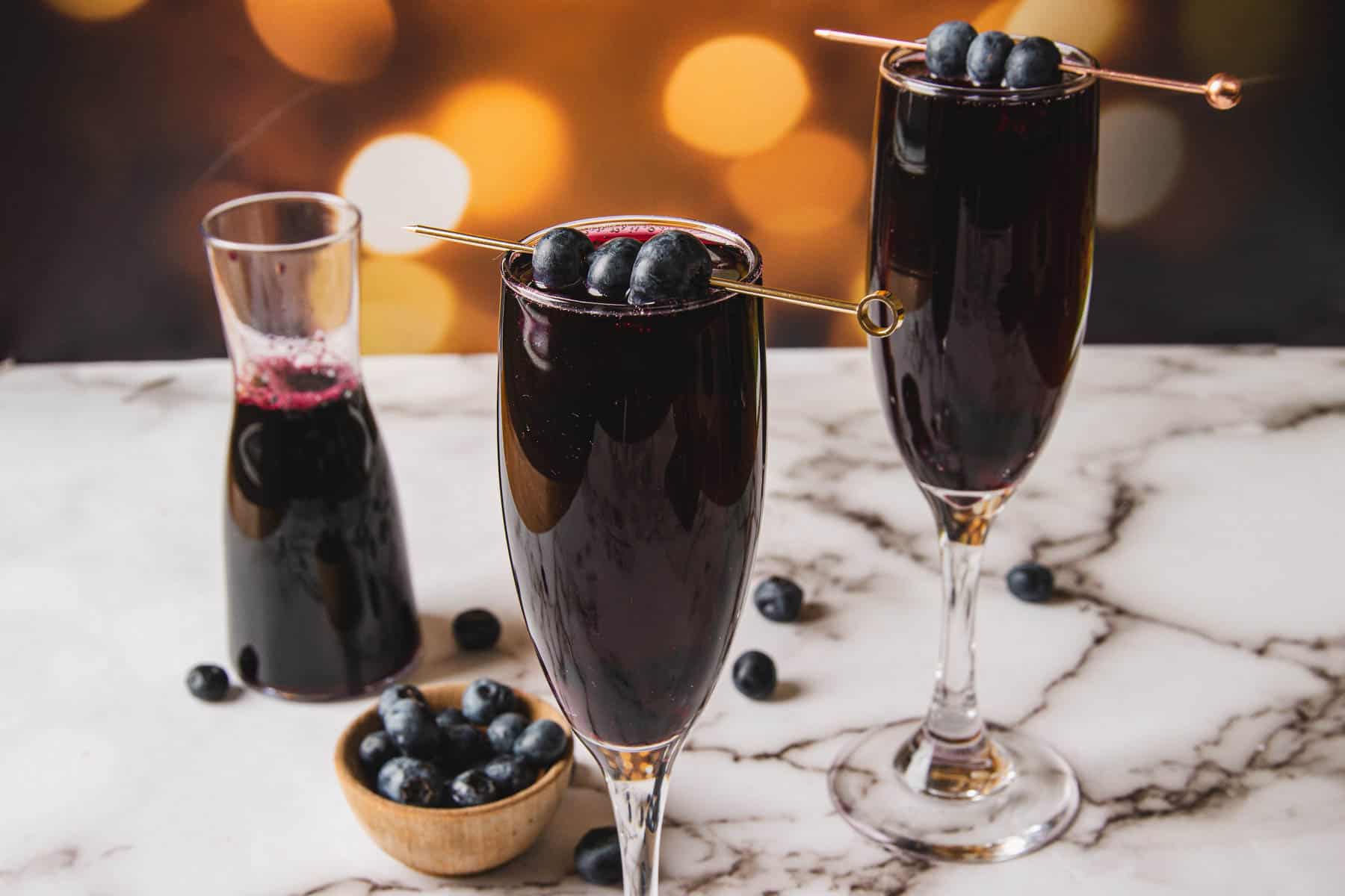 Two glasses of dark red liquid, reminiscent of a Kir Royale, are elegantly garnished with blueberries on skewers on a marble surface. Nearby, a small bowl of blueberries and a pitcher brim with the same tantalizing beverage, set against a bokeh background in warm tones.