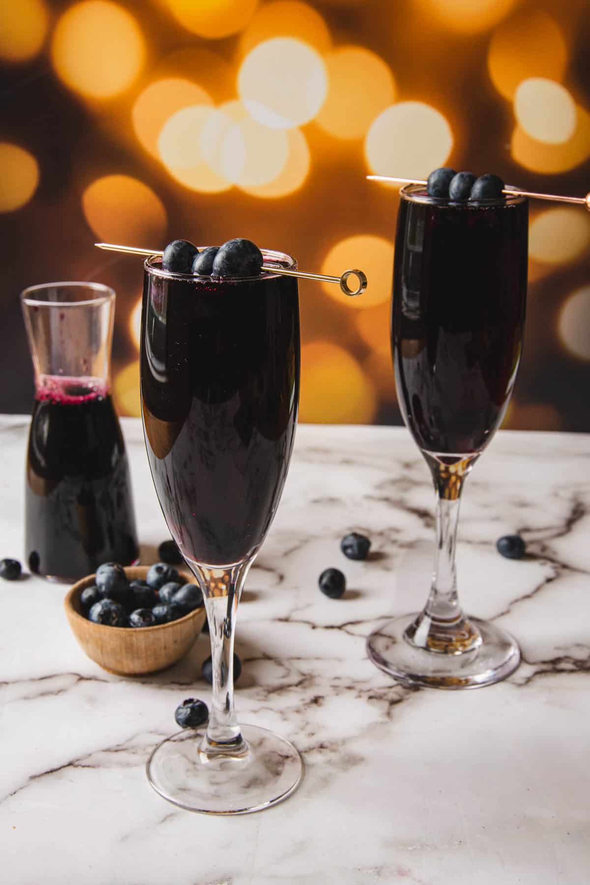 Two champagne flutes, each filled with a rich blend resembling kir royale, are garnished with skewered blueberries on a marble surface. A small bowl of blueberries and a carafe sit in the background against a warm, blurred light backdrop.