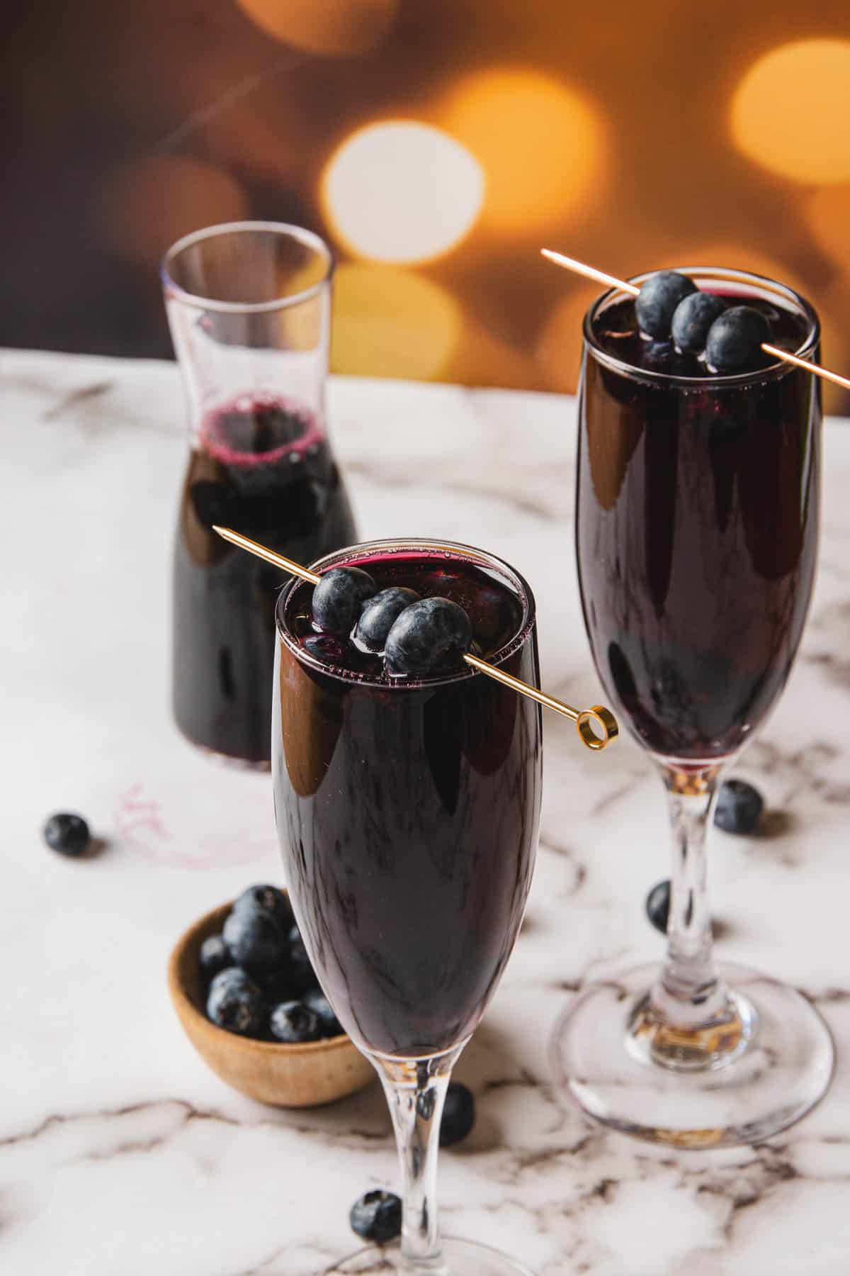 Two glasses of a dark beverage, reminiscent of a Kir Royale's elegance, are adorned with skewered blueberries on a marble surface. In the background, a small wooden bowl brims with blueberries next to a carafe filled with the same enticing drink.