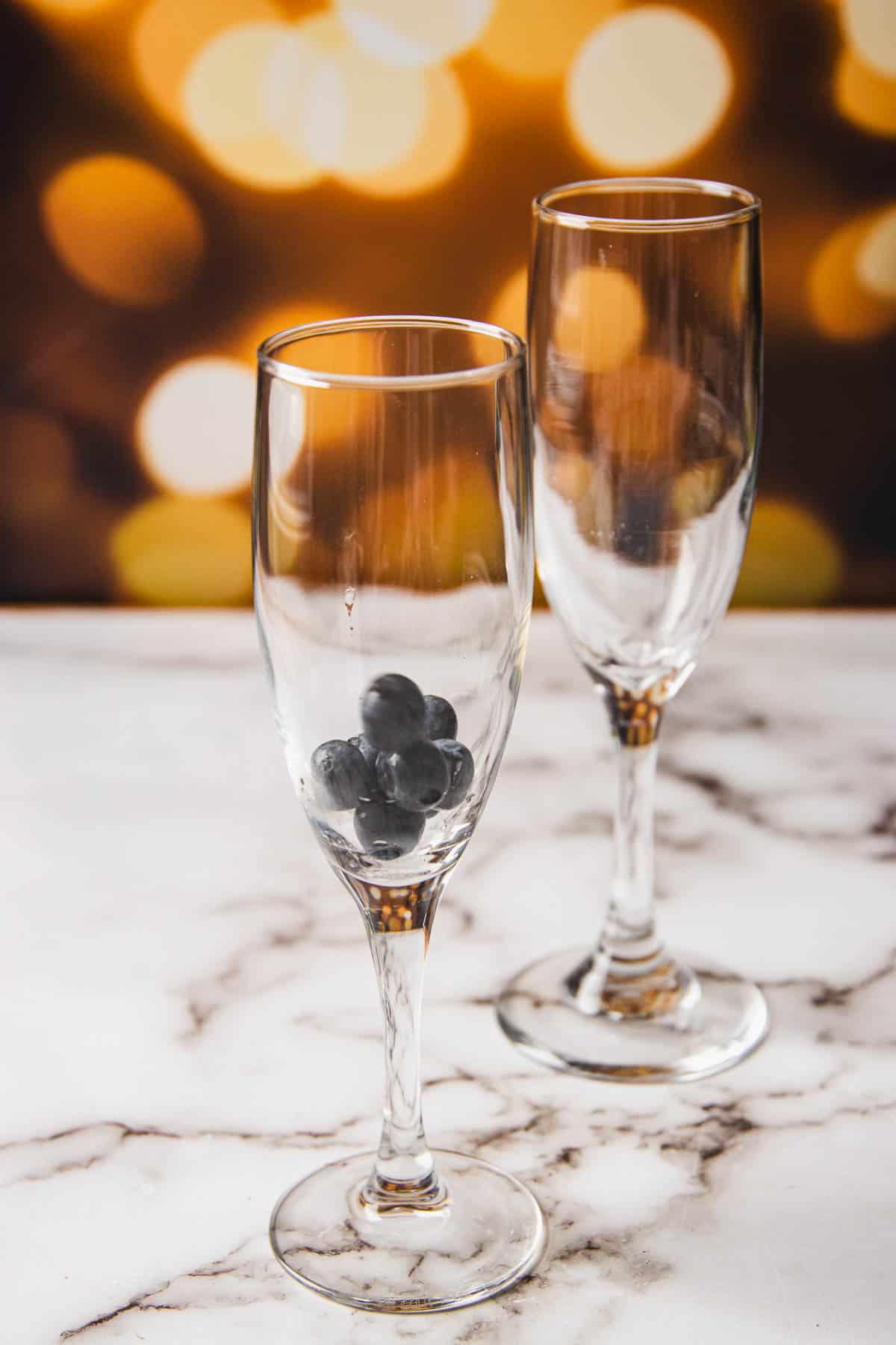 Two empty champagne glasses with a few blueberries inside sit on a marble surface, offering a nod to what is Kir Royale. A warm, blurred background with circular bokeh lights adds a decorative touch.