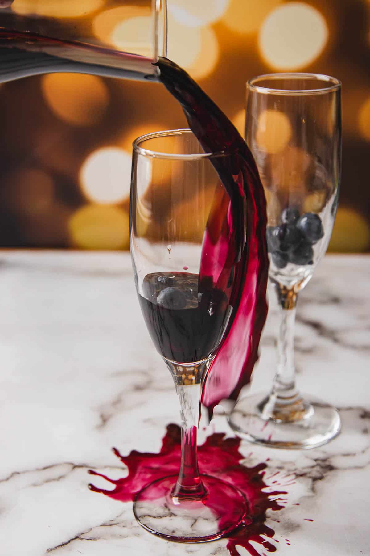 Red wine is being poured into a wine glass, overflowing and spilling onto the marble surface. Another wine glass with a few blueberries inside sits nearby, reminiscent of a kir royale. In the background, blurred golden lights create a cozy ambiance.