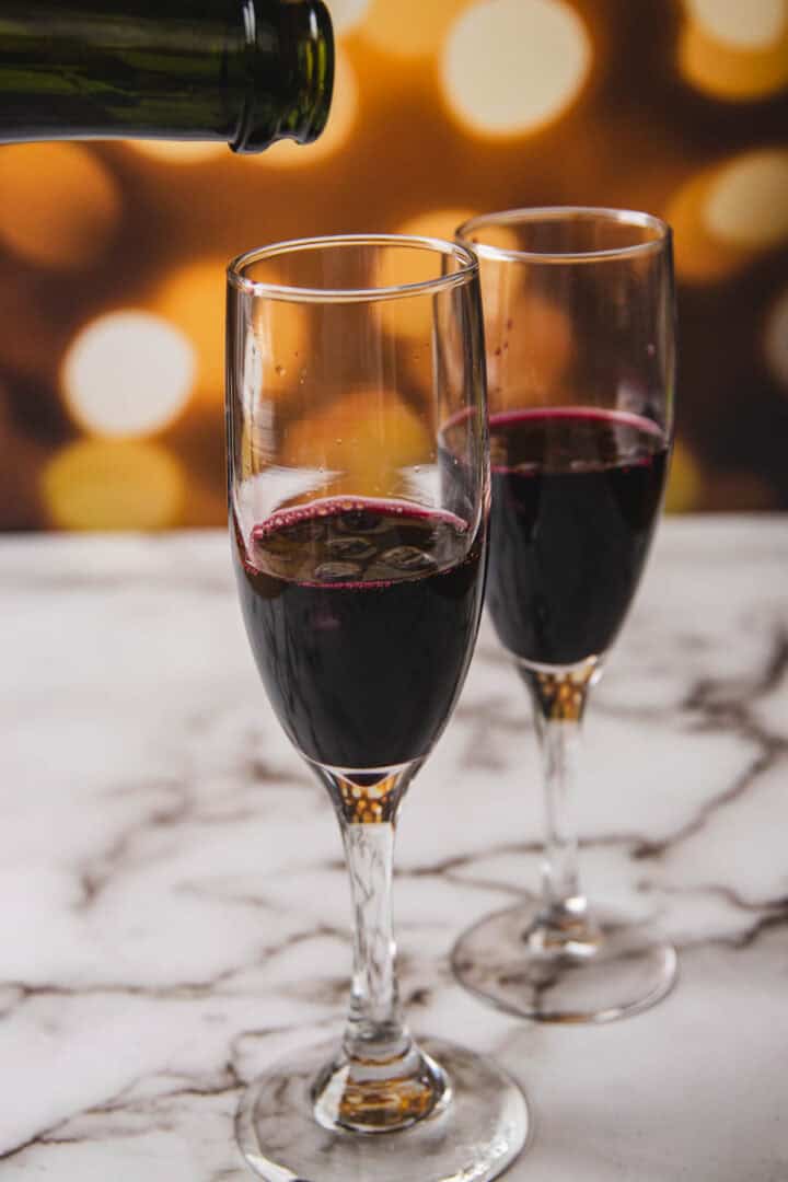 Two champagne flutes rest on a marble surface as red wine is poured into one, evoking the elegance of a Kir Royale. The background features blurred circular lights, creating a warm ambiance.