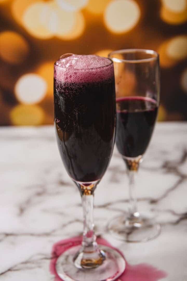 Two champagne flutes are filled with a dark red sparkling liquid, reminiscent of a classic Kir Royale. The glass in the foreground has a bubbly foam at the top with some liquid spilled around its base. In the background, blurry circular lights create a festive ambiance.