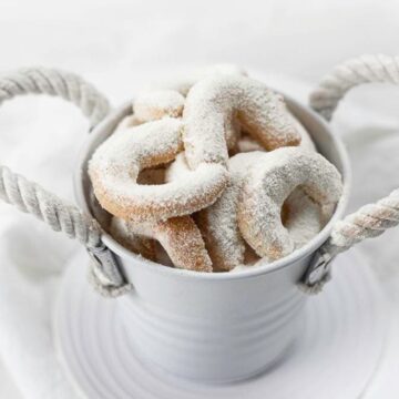 A white metal bucket with rope handles holds irresistible sugar-dusted Christmas cookies, beautifully crescent-shaped. The bucket sits elegantly on a white surface.