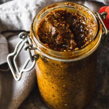 A jar filled with a chunky, brown paste sits on a dark surface, ready to be the show-stealing dip for your chips. Beside it rests a folded gray cloth. The jar features a metal clasp and a striking red lid, adding charm to any holiday spread.