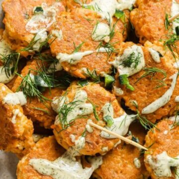 A plate of orange-hued salmon fritters topped with a creamy dill sauce and garnished with fresh dill sprigs. The fritters are round and have visible chunks of salmon and herbs, arranged neatly and pierced with wooden skewers.