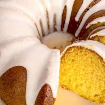 A close-up of a Bundt cake on a yellow plate. The cake has a golden-brown crust and is topped with white icing. A slice has been cut, revealing the moist interior. A blue and white checkered cloth is partially visible in the background.