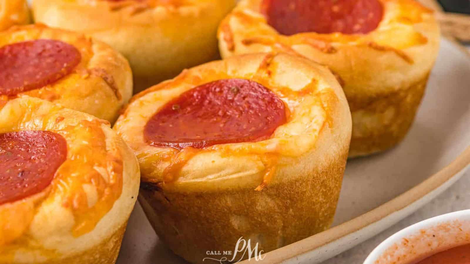 Close-up of several pizza muffins on a plate, each topped with melted cheese and a slice of pepperoni. With their golden-brown crust and circular arrangement, these kid-friendly delights are sure to please even the pickiest eaters. A small portion of a wooden utensil is visible on the side.