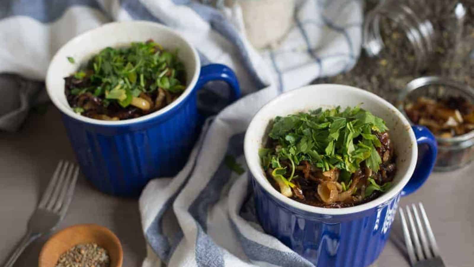 Two blue ceramic mugs filled with cooked vegetables and topped with chopped herbs are placed on a table. A striped cloth and scattered dried ingredients are nearby, along with forks and a small bowl of mixed seeds.